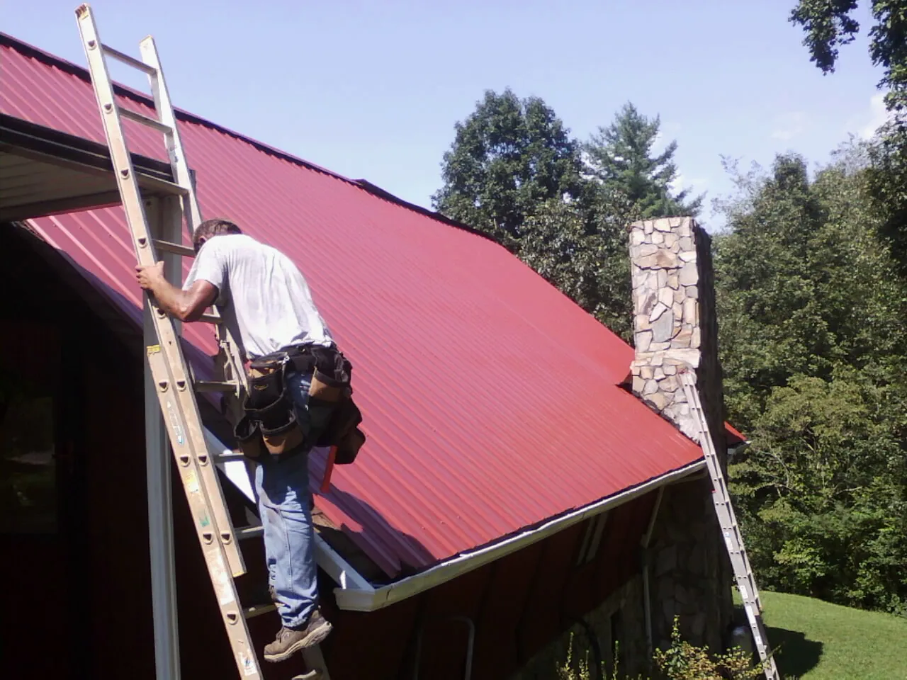 Roofer on a ladder beside a red metal roof and stone chimney in Breesport